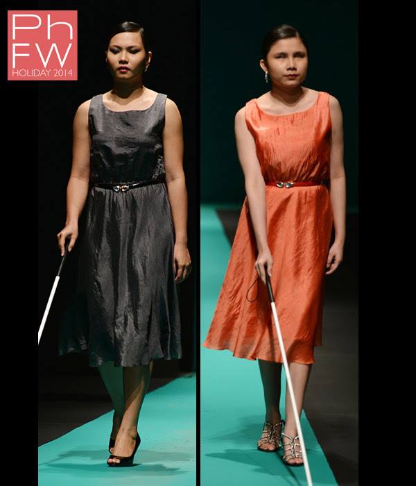 Two models walking on a runway at PhFW Holiday 2014. The left model is wearing a sleeveless black dress with a belt and holding a white cane. The right model is in an orange sleeveless dress, also with a belt, and is holding a white cane. Both have dark hair styled neatly and wear heeled shoes. The background is dark, with a teal runway floor.